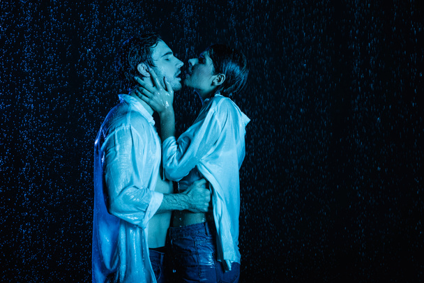 sexy pareja romántica húmeda suavemente abrazos y besos en gotas de agua sobre fondo negro con filtro de color azul - Foto, imagen