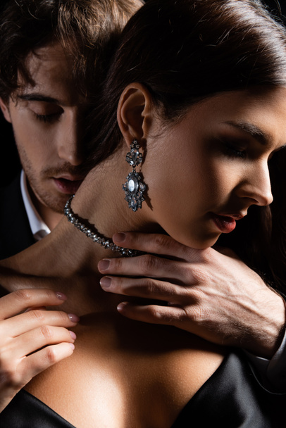 close up shot of man passionately holding hands on neck of woman in silk dress and jewelry isolated on black - Photo, Image
