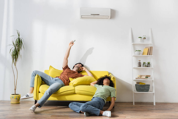 young man holding remote controller near air conditioner while suffering from heat with african american girlfriend - 写真・画像