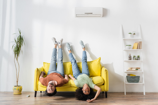 junges interrassisches Paar leidet unter Hitze auf gelbem Sofa im modernen Wohnzimmer  - Foto, Bild