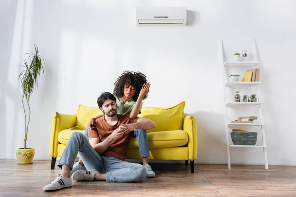 Unzufriedenes gemischtrassiges Paar blickt auf Fernbedienung, während es auf Couch in der Nähe von Klimaanlage sitzt  - Foto, Bild