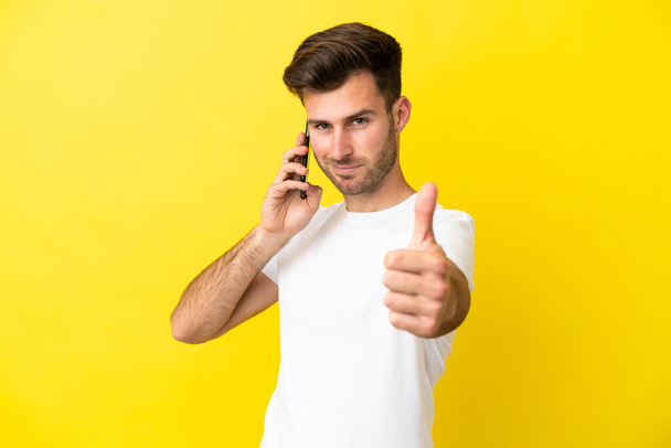 Молодий кавказький красивий чоловік ізольований на жовтому тлі, тримаючи розмову з мобільним при цьому роблячи великі пальці вгору
 - Фото, зображення