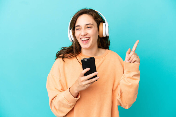 Νεαρή καυκάσια γυναίκα απομονωμένη σε μπλε φόντο ακούγοντας μουσική με κινητό και τραγουδώντας - Φωτογραφία, εικόνα