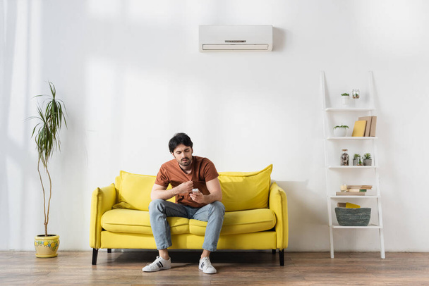 Mann hält Fernbedienung in der Hand, während er unter Hitze im Wohnzimmer leidet - Foto, Bild