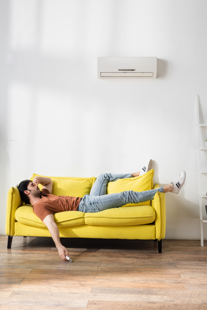 hombre sosteniendo mando a distancia mientras está acostado en el sofá amarillo y que sufre de calor en la sala de estar - Foto, Imagen