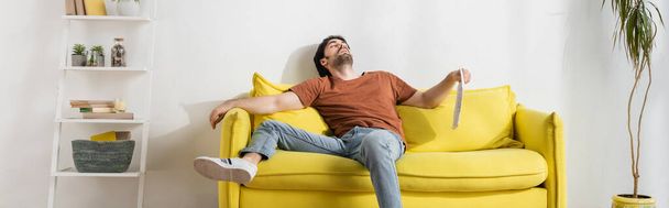 hombre sosteniendo periódico mientras está acostado en el sofá amarillo y sufriendo de calor en la sala de estar, pancarta - Foto, imagen