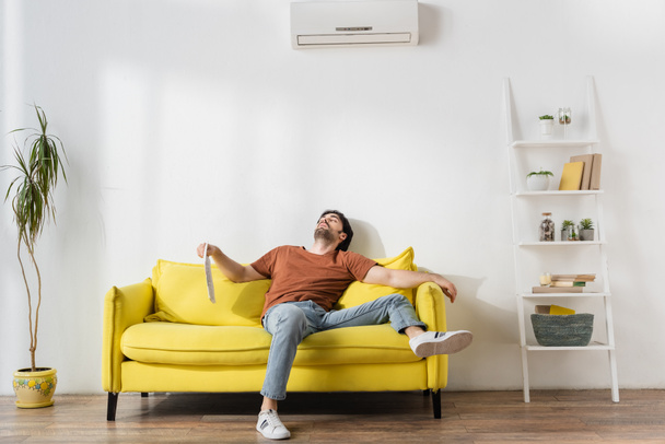 hombre sosteniendo el periódico mientras está acostado en el sofá amarillo y sufriendo de calor en la sala de estar - Foto, Imagen