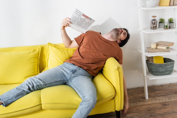 man waving with newspaper while lying on yellow couch and suffering from heat - Φωτογραφία, εικόνα