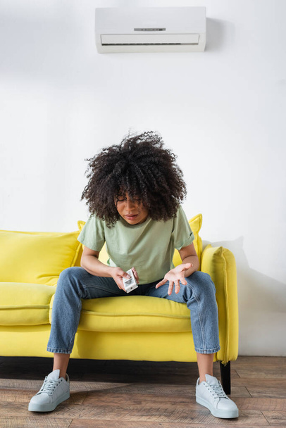 displeased african american woman holding remote controller while sitting on yellow couch and suffering from heat - Foto, Bild