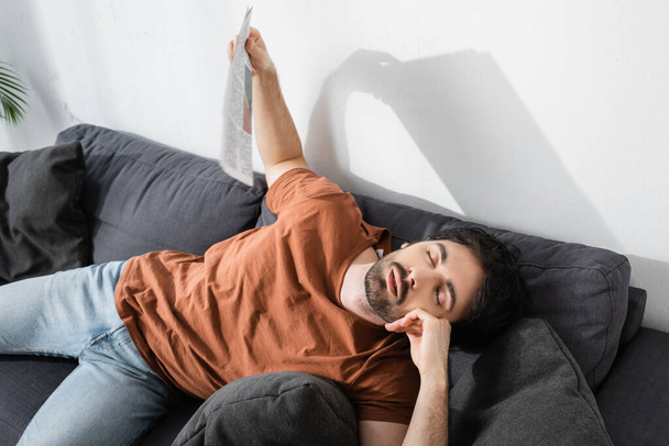 vista de ángulo alto del hombre con los ojos cerrados ondeando con el periódico mientras está acostado en el sofá gris y sufriendo de calor - Foto, imagen