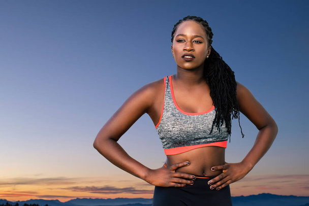 Zbliżenie portret młodej atrakcyjnej afrykańskiej nastolatki atletki stojącej z rękami na biodrach. Średni strzał muskularnej dziewczyny o zachodzie słońca przeciwko ciemnemu błękitnemu niebu i górom. - Zdjęcie, obraz