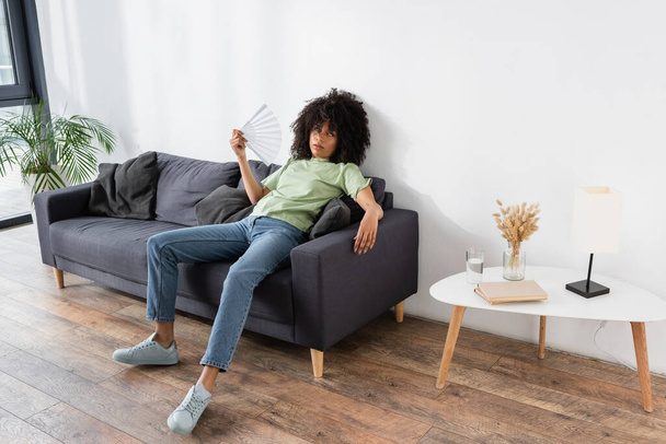 african american woman waving with hand fan while sitting on grey couch and suffering from heat - Foto, Bild