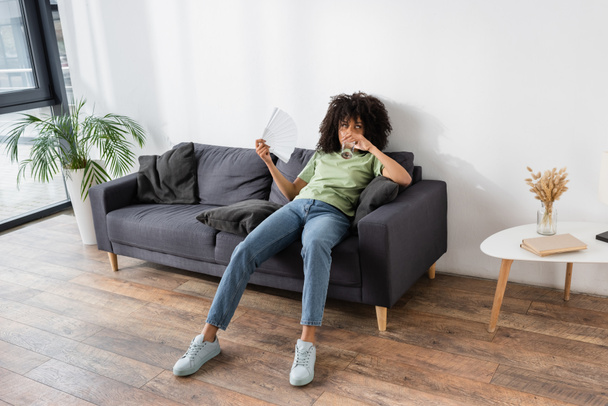 african american woman waving with hand fan while drinking water and sitting on grey couch  - Foto, Bild