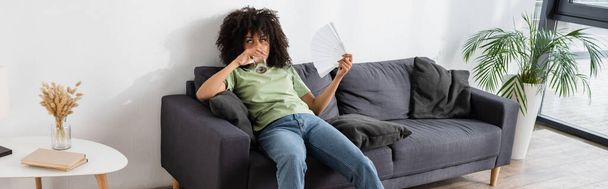 donna afroamericana che sventola con ventilatore a mano mentre beve acqua e siede sul divano grigio, striscione - Foto, immagini