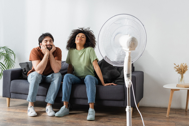 potěšený mezirasový muž a žena sedí na gauči v blízkosti rozmazané elektrický ventilátor v obývacím pokoji  - Fotografie, Obrázek