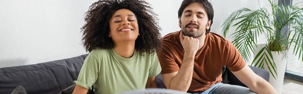 happy interracial couple sitting on couch in living room, banner - Foto, imagen