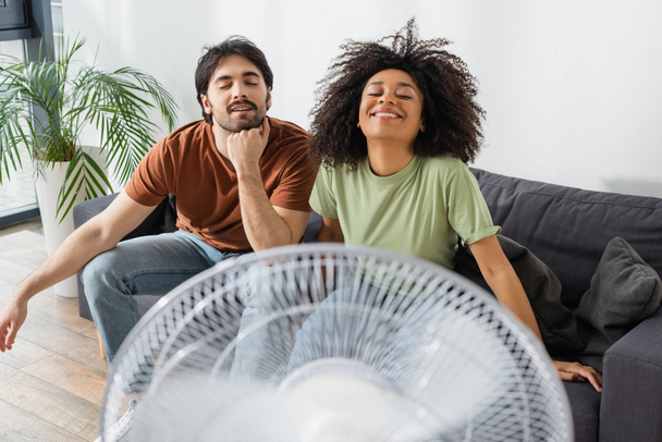 joyful interracial couple sitting on couch near blurred electric fan in living room  - Photo, Image
