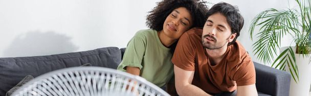 pleased interracial couple sitting on couch near blurred electric fan in living room, banner  - Fotó, kép