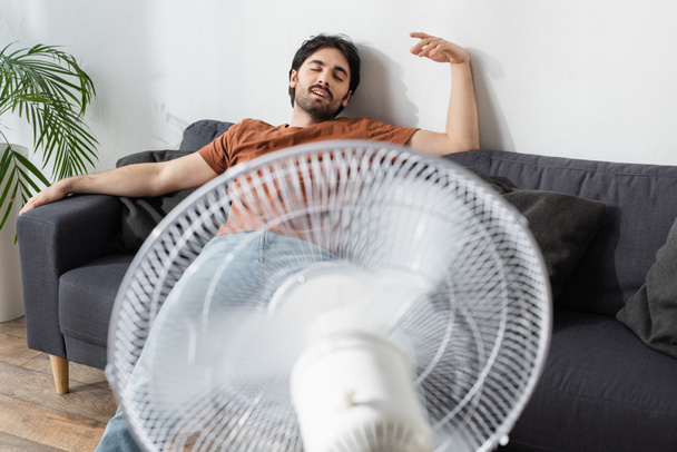 pleased bearded man sitting on couch near blurred electric fan  - Fotó, kép