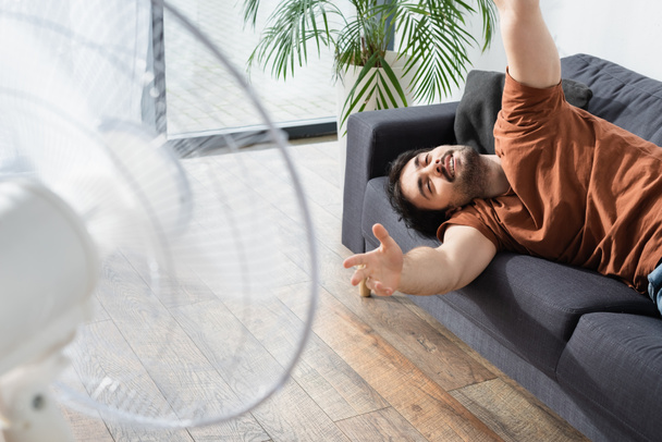 довольный бородатый мужчина лежит на диване рядом с размытым электрическим вентилятором  - Фото, изображение