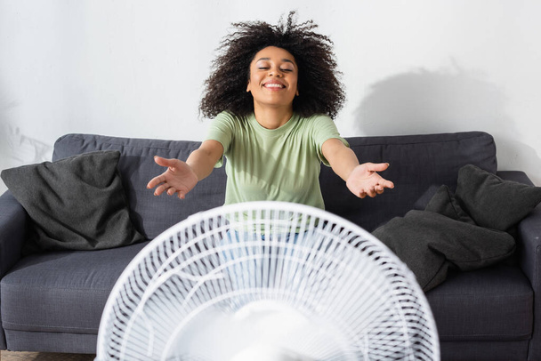 Радостная африканская американка сидит с протянутыми руками на диване рядом с размытым электрическим вентилятором - Фото, изображение