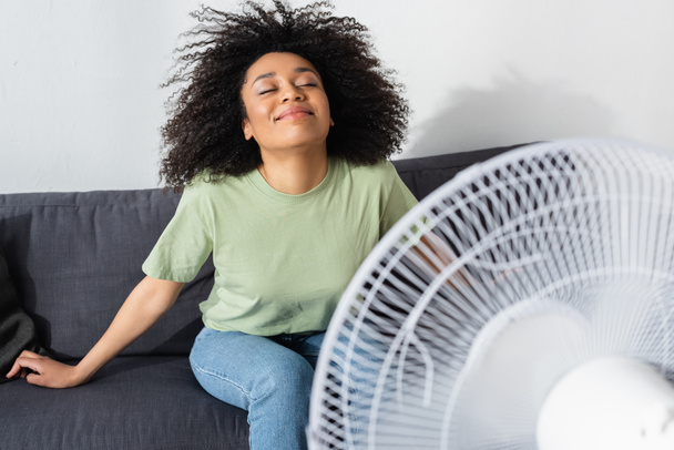 joyeuse femme afro-américaine assise sur le canapé près d'un ventilateur électrique flou - Photo, image