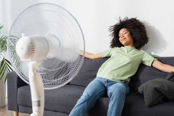 pleased african american woman resting on couch near blurred electric fan - Foto, afbeelding