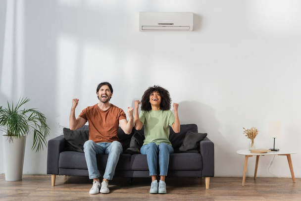happy multiethnic rejoicing while sitting on couch in modern living room, hvac concept  - Photo, Image