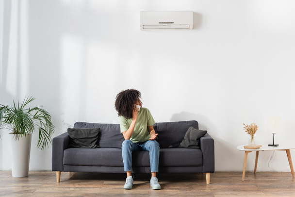 mulher afro-americana descontente falando no smartphone e olhando para o ar condicionado na sala de estar  - Foto, Imagem