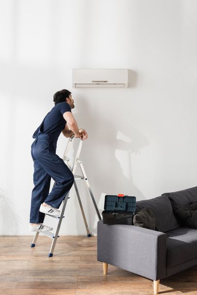 bearded handyman in overalls standing on ladder and looking at broken air conditioner  - Φωτογραφία, εικόνα