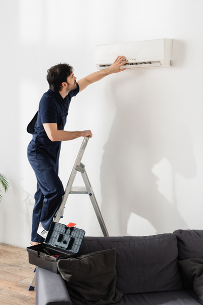 bearded handyman in overalls standing on ladder and fixing broken air conditioner  - Photo, Image