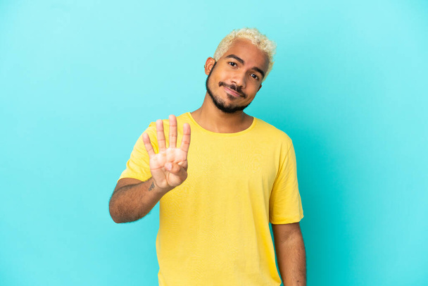 młody Kolumbijczyk przystojny mężczyzna odizolowany na niebieskim tle szczęśliwy i liczyć cztery palce - Zdjęcie, obraz