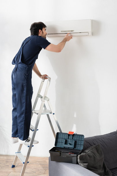 young handyman in overalls standing on ladder while fixing broken air conditioner  - Photo, Image