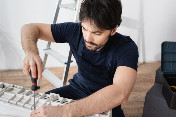 bearded handyman holding screwdriver while fixing broken air conditioner  - Valokuva, kuva