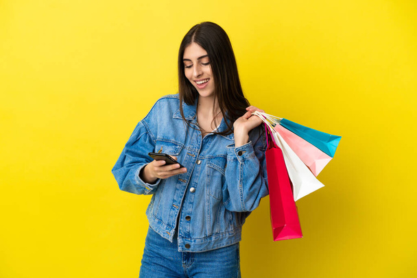 Nuori valkoihoinen nainen eristyksissä sinisellä taustalla pitäen ostoskasseja ja kirjoittaen viestiä matkapuhelimellaan ystävälleen. - Valokuva, kuva