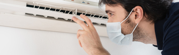 handyman in medical mask gesturing near air conditioner while checking climate control, banner - Фото, изображение