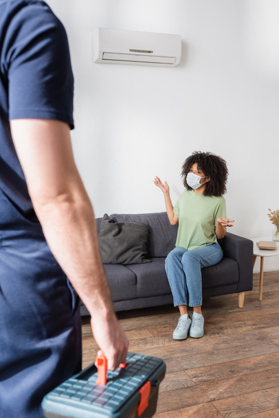 african american woman in medical mask pointing at broken air conditioner near blurred handyman with toolbox  - Photo, Image