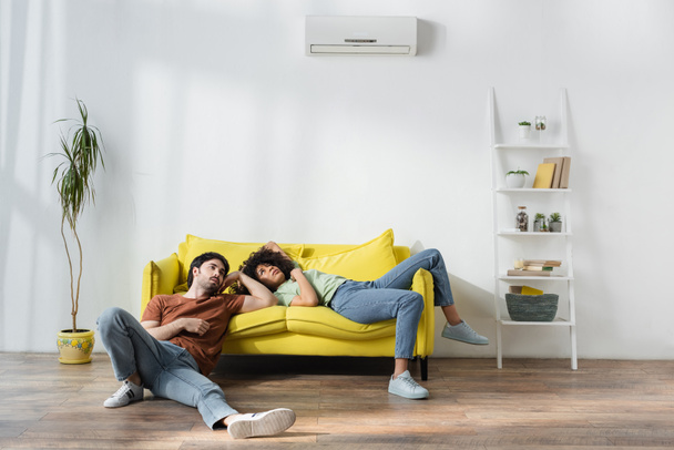 tired multiethnic couple sitting on yellow sofa and suffering from heat in summer - Photo, Image