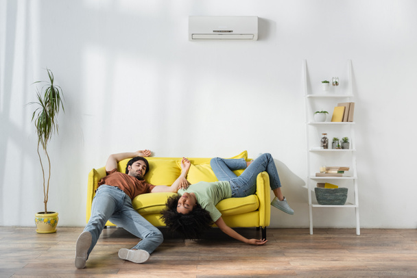 épuisé jeune couple multiethnique couché sur un canapé jaune et souffrant de chaleur en été - Photo, image