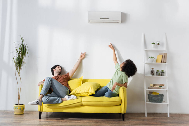 multiethnic couple sitting on yellow sofa and reaching air conditioner while suffering from heat in summer - Photo, Image