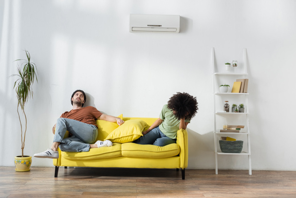 pareja interracial agotada sentado en el sofá y sufriendo de calor en verano - Foto, imagen