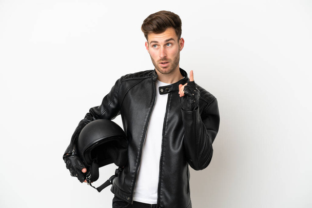 Молодой кавказский человек в мотоциклетном шлеме на белом фоне думает об идее, указывая пальцем вверх - Фото, изображение