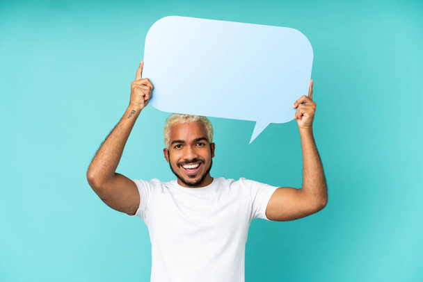 Young Colombian handsome man isolated on blue background holding an empty speech bubble - Фото, изображение