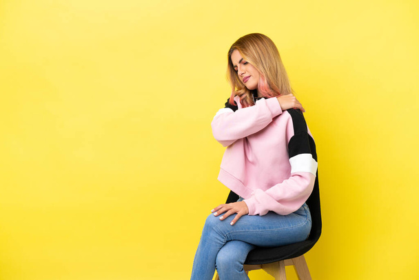 Молода жінка сидить на стільці над ізольованим жовтим тлом, страждає від болю в плечі за те, що зробила зусилля
 - Фото, зображення