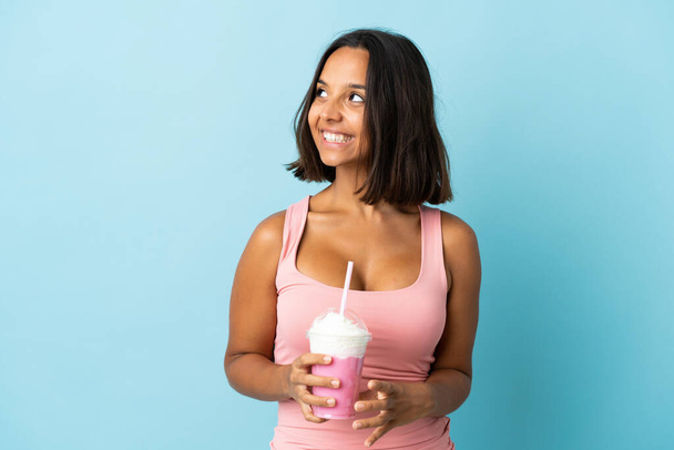 jonge vrouw met aardbei milkshake geïsoleerd op blauwe achtergrond denken een idee terwijl omhoog kijken - Foto, afbeelding