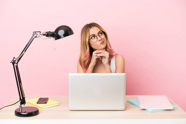 Молода студентка на робочому місці з ноутбуком на рожевому фоні має сумніви під час пошуку
 - Фото, зображення