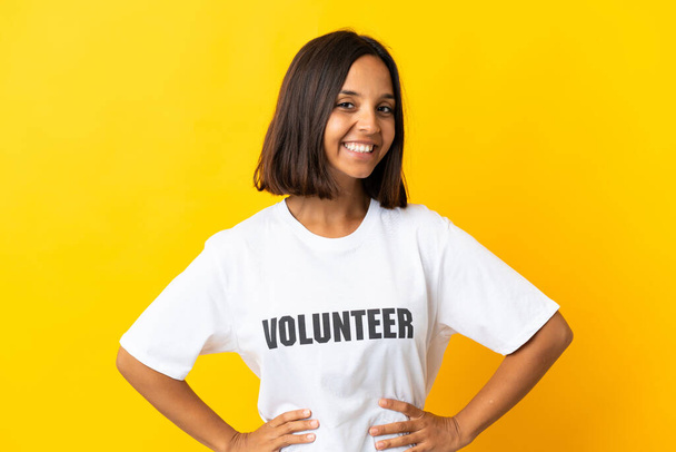 Young volunteer woman isolated on yellow background posing with arms at hip and smiling - Foto, Imagen