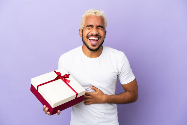 Young Colombian man holding a gift isolated on purple background smiling a lot - Фото, изображение