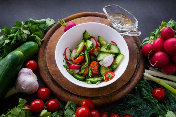 A pile of fresh, ripe vegetables: radishes, tomatoes, cucumbers, herbs, and garlic are arranged around a round wooden chopping board. A white bowl with vegetables cut into a salad and olive oil. - Photo, Image