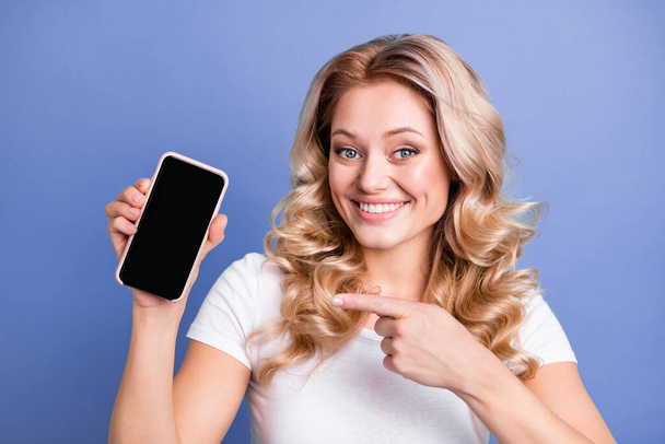 Portrait of attractive cheerful wavy-haired girl holding in hand demonstrating device screen isolated over blue color background - Φωτογραφία, εικόνα